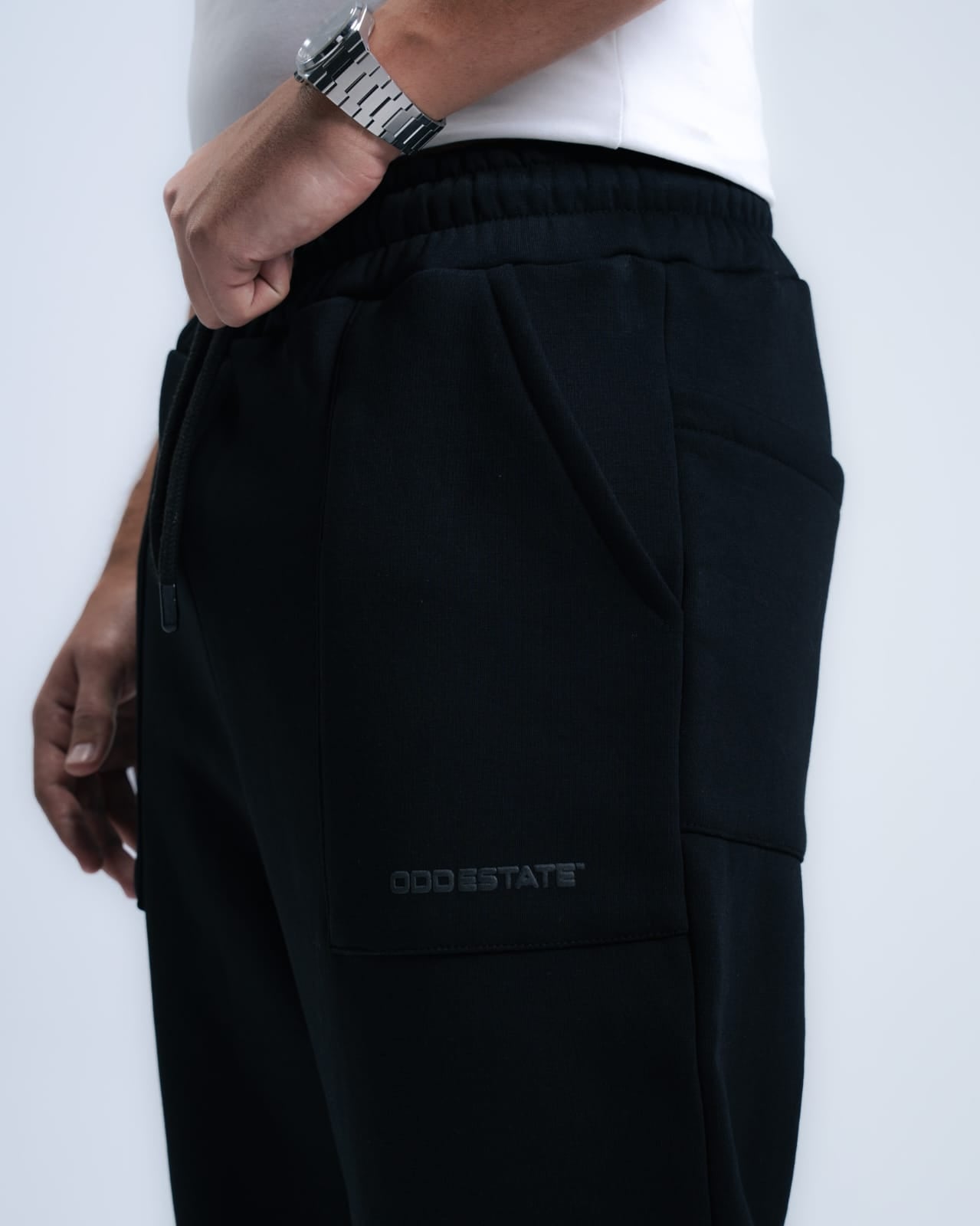 A black baggy sweatpants and white t-shirt with a light grey background with a metal watch